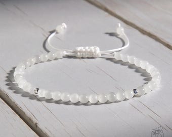 May include: White beaded bracelet with a macrame fastening and small silver accents. The bracelet features small, round, white beads strung on a white cord. The macrame fastening allows for adjustable sizing. The bracelet is on a light-coloured wooden surface.