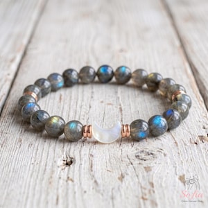 May include: A beaded bracelet featuring round, grey labradorite stones with iridescent blue and gold flashes. The bracelet includes a central white crescent moon charm and small rose gold-toned metal spacers between the beads.