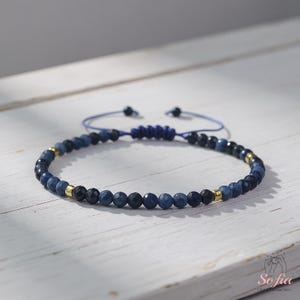 May include: A beaded bracelet featuring a combination of dark blue and black beads, interspersed with small gold-colored accents. The bracelet is strung on a blue cord with an adjustable sliding knot closure. The bracelet is displayed on a white wooden surface.