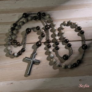 K&ouml;nnte beinhalten: Ein Rosenkranz mit einem silbernen Kreuzanh&auml;nger und Perlen in Grau- und Dunkelbraunt&ouml;nen. Die Perlen sind auf einer silbernen Kette aufgef&auml;delt. Der Rosenkranz wird auf einer hellen Holzoberfl&auml;che pr&auml;sentiert.