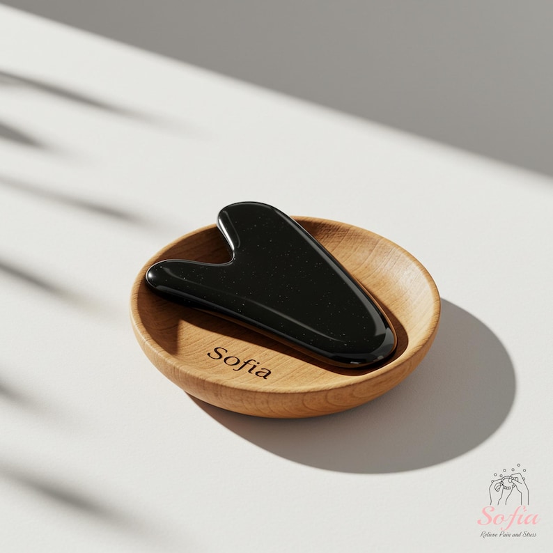 May include: A black gua sha tool sits in a small wooden bowl. The bowl is engraved with the name "Sofia". The gua sha is smooth and shiny, and the bowl is light brown. The background is white with shadows.