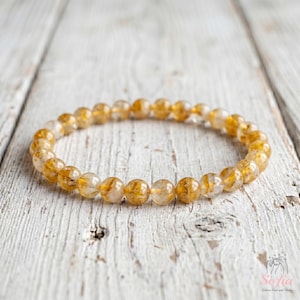 May include: A circular stretch bracelet composed of numerous translucent, spherical beads. Each bead features internal golden-yellow inclusions that create a shimmering, fibrous appearance within the clear, polished stone material.