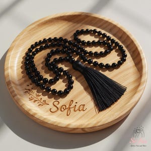 May include: A black beaded necklace with a tassel, displayed on a wooden tray. The tray is engraved with the name "Sofia" and a floral design. The necklace is made of small, round beads.