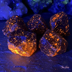 May include: Five dark, rough-textured, irregularly shaped stones with orange flecks, glowing under a UV light.