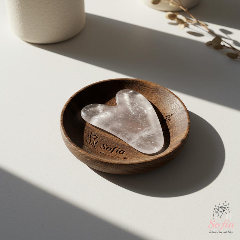 May include: A heart-shaped, clear quartz gua sha tool sits in a small, round wooden bowl. The bowl is engraved with the word "Sofia" and a floral design. The gua sha is on a white surface, with shadows.