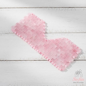 May include: A pink rose quartz eye mask made from small, rectangular stones. The mask is edged with small, round beads and is designed to cover the eyes. The mask is on a white wooden surface.