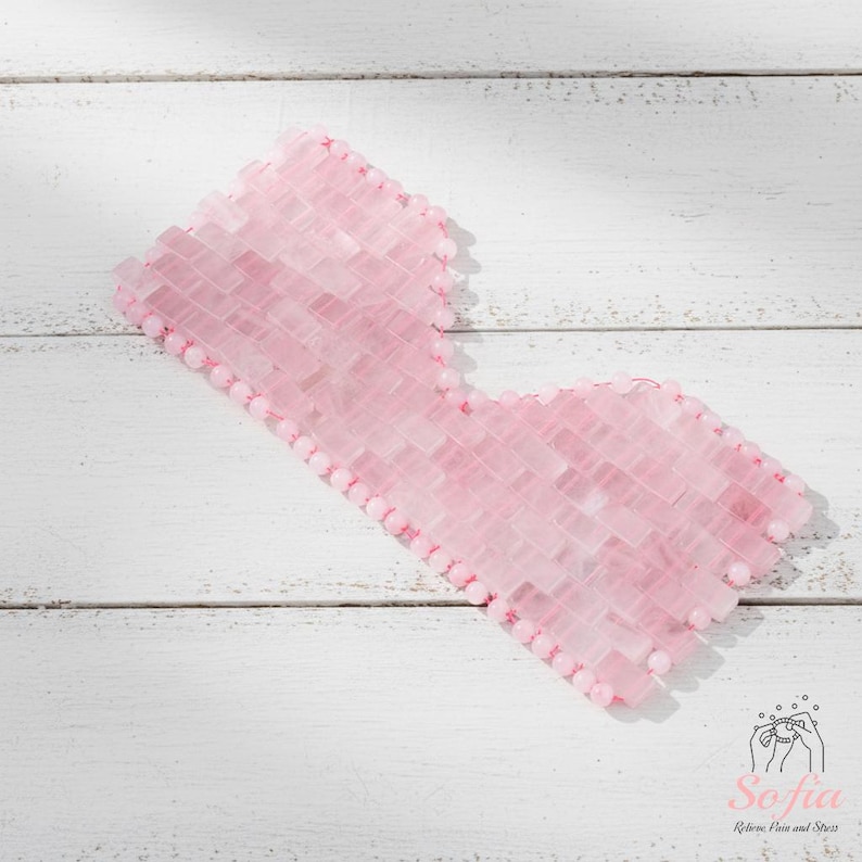 May include: A pink rose quartz eye mask made from small, rectangular stones. The mask is edged with small, round beads and is designed to cover the eyes. The mask is on a white wooden surface.