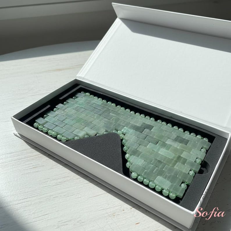 May include: A jade eye mask made of small, rectangular jade stones, displayed in a white box with a black interior. The mask is light green and has a beaded edge. The box is open, showing the mask.