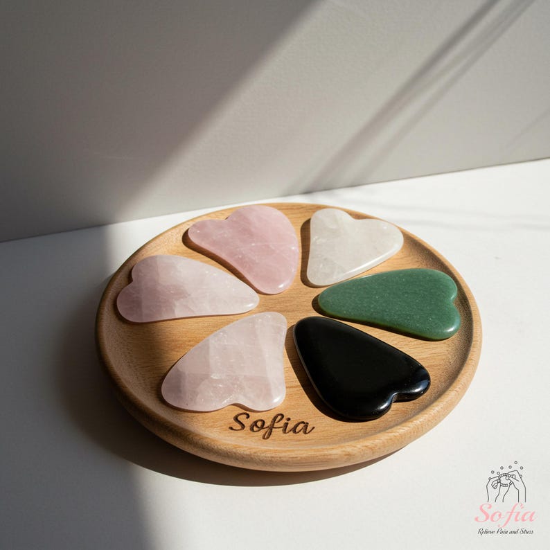 May include: A wooden tray displaying seven heart-shaped gua sha tools in shades of pink, green, white, and black. The tray is engraved with the name "Sofia".