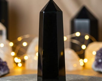 May include: A black obsidian crystal point, also known as a black obelisk, is a polished, faceted, and pointed crystal. The crystal is on a gray surface.