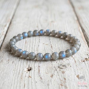 May include: A circular beaded bracelet composed of smooth, round labradorite stones. Each bead displays a grey base color with iridescent blue flashes that catch the light, arranged in a continuous loop on an elastic cord for a flexible fit.