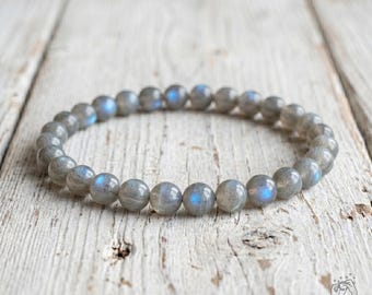 May include: A circular beaded bracelet composed of smooth, round labradorite stones. Each bead displays a grey base color with iridescent blue flashes that catch the light, arranged in a continuous loop on an elastic cord for a flexible fit.