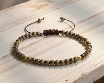 May include: A beaded bracelet featuring faceted, metallic-gold beads strung on a brown cord. The adjustable bracelet has a sliding knot closure and is displayed on a light-colored wooden surface.