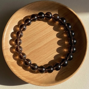May include: A circular bracelet composed of translucent, dark brown, spherical beads strung together. The beads have a smooth, polished finish and exhibit a deep, smoky color that allows light to pass through, creating subtle highlights on their surfaces.