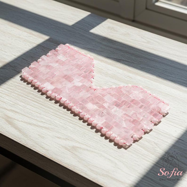 May include: A pink rose quartz eye mask made of small, rectangular stones. The mask is designed to cover the eyes and part of the cheeks. The stones are strung together with a pink thread. The mask is on a light wood surface.