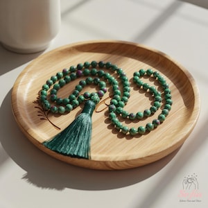 May include: A green beaded necklace with a tassel, presented on a wooden tray. The necklace features green beads with red accents and a matching green tassel. The wooden tray has a natural wood grain finish.