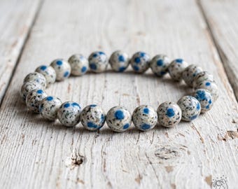 May include: A beaded bracelet composed of round, polished stones featuring a speckled cream and gray base with distinct, vibrant blue circular spots. The beads are strung together in a circular formation, creating a uniform, elasticized accessory.