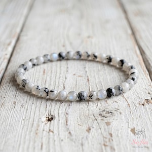 Op de afbeelding: Een kralenarmband bestaande uit kleine, ronde, doorschijnende witte stenen met verspreide zwarte insluitingen en subtiele iriserende blauwe flitsen. De kralen zijn in een cirkelvorm geregen, wat een delicaat en uniform accessoire vormt.