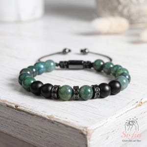 May include: A beaded bracelet featuring alternating matte black spheres and polished green mottled stone beads. The design includes two dark metal spacers encrusted with small black crystals and a black adjustable cord closure.