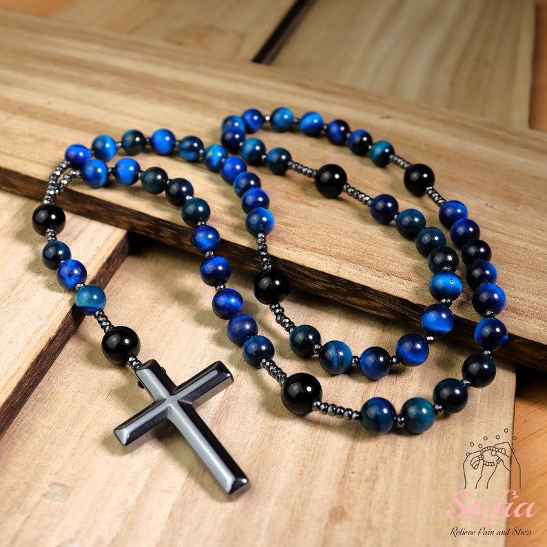 Pu&ograve; includere: Un rosario con perline blu e nere e un grande ciondolo a croce in metallo nero. Le perline sono in occhio di tigre e ematite.
