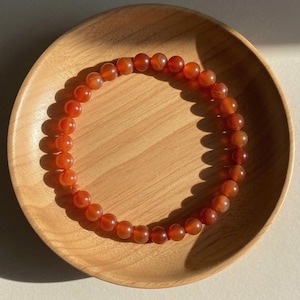 May include: A circular beaded bracelet composed of numerous smooth, polished, translucent orange-red carnelian gemstone spheres. The beads are uniform in size and strung together in a continuous loop, displaying natural variations in color intensity.
