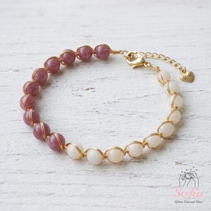 May include: A bracelet featuring a series of round mauve and white beads, each individually wrapped in thin gold-tone wire. The piece includes a gold-tone lobster claw clasp, an adjustable chain, and a small heart-shaped charm at the end.