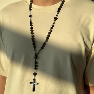 K&ouml;nnte beinhalten: Eine lange Perlenkette mit einem Kreuzanh&auml;nger. Die Kette hat dunkelgraue und schwarze Perlen mit einem schwarzen Kreuz am unteren Ende. Die Kette wird &uuml;ber einem cremefarbenen T-Shirt getragen.