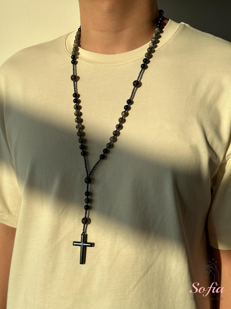 K&ouml;nnte beinhalten: Eine lange Perlenkette mit einem Kreuzanh&auml;nger. Die Kette hat dunkelgraue und schwarze Perlen mit einem schwarzen Kreuz am unteren Ende. Die Kette wird &uuml;ber einem cremefarbenen T-Shirt getragen.