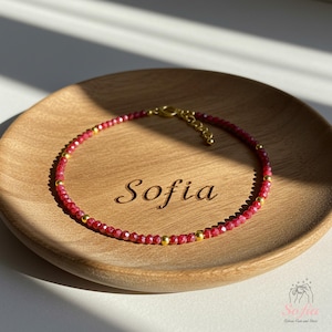 May include: A red gemstone and gold bead bracelet with a clasp. The bracelet is on a wooden surface with the name "Sofia" engraved on it.