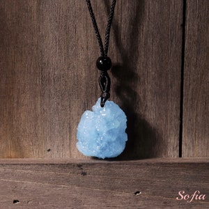 Puede incluir: Un colgante de cristal azul claro en un collar de cord&oacute;n negro. El colgante tiene una forma irregular y rugosa y un acabado brillante y brillante.