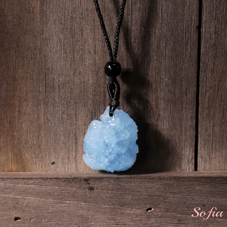 Puede incluir: Un colgante de cristal azul claro en un collar de cord&oacute;n negro. El colgante tiene una forma irregular y rugosa y un acabado brillante y brillante.