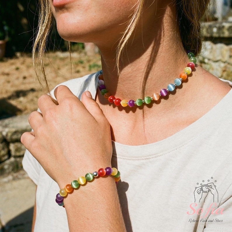 May include: A necklace and bracelet set featuring round, multicolored cat's eye beads in shades of red, orange, yellow, green, blue, and purple. The beads are arranged in a repeating rainbow sequence, separated by small silver-toned metal spacer beads.
