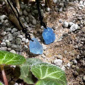Puede incluir: Dos colgantes de cristal azul claro en cordones negros. Los cristales son rugosos y tienen una forma natural irregular. Los cordones est&aacute;n atados con cuentas negras.