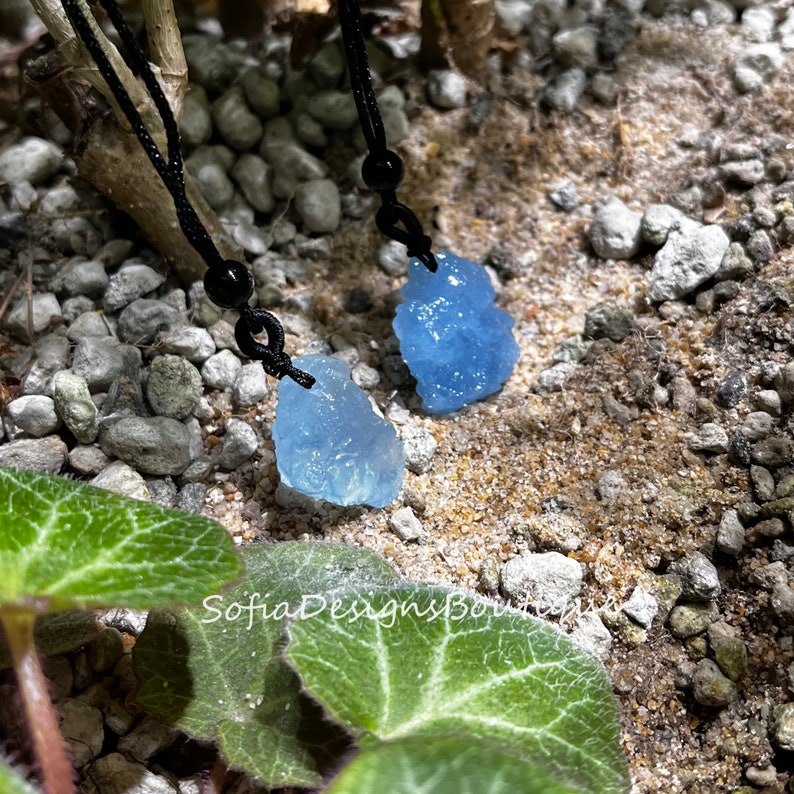Puede incluir: Dos colgantes de cristal azul claro en cordones negros. Los cristales son rugosos y tienen una forma natural irregular. Los cordones est&aacute;n atados con cuentas negras.
