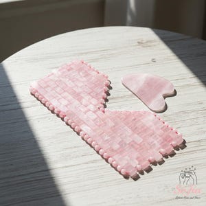 May include: A pink rose quartz eye mask and a gua sha tool rest on a white wooden surface. The eye mask is constructed from small, rectangular rose quartz pieces, and the gua sha tool is heart-shaped. These items are designed for skincare and relaxation, commonly used in beauty routines.