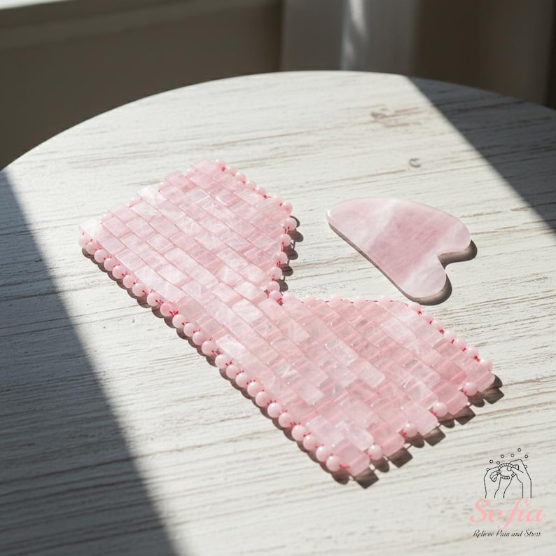 May include: A pink rose quartz eye mask and a gua sha tool rest on a white wooden surface. The eye mask is constructed from small, rectangular rose quartz pieces, and the gua sha tool is heart-shaped. These items are designed for skincare and relaxation, commonly used in beauty routines.