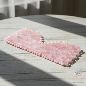 May include: A pink rose quartz eye mask with a rectangular shape and rounded edges. The mask is made of small, rectangular pieces of rose quartz and is surrounded by a row of small, round beads. The mask is designed to relieve pain and stress.