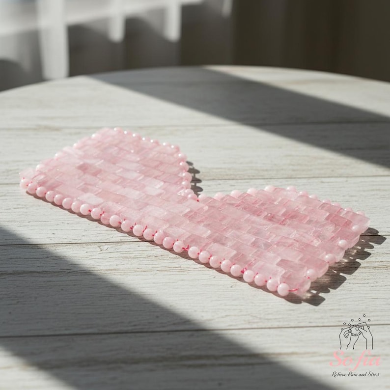 May include: A pink rose quartz eye mask with a rectangular shape and rounded edges. The mask is made of small, rectangular pieces of rose quartz and is surrounded by a row of small, round beads. The mask is designed to relieve pain and stress.
