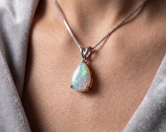 Könnte beinhalten: Ein tropfenförmiger Opal-Anhänger in einer silberfarbenen Metallfassung, der an einer zarten silberfarbenen Box-Kette hängt. Der Opal zeigt ein lebhaftes Farbspiel mit roten, grünen und blauen Lichtreflexen auf einem weißen Untergrund.