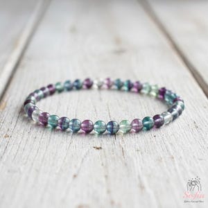 May include: A beaded bracelet featuring small, round, polished fluorite beads in alternating shades of translucent purple, deep violet, and soft seafoam green. The beads are strung together in a continuous circular loop, creating a smooth, uniform appearance throughout the entire piece of jewelry.