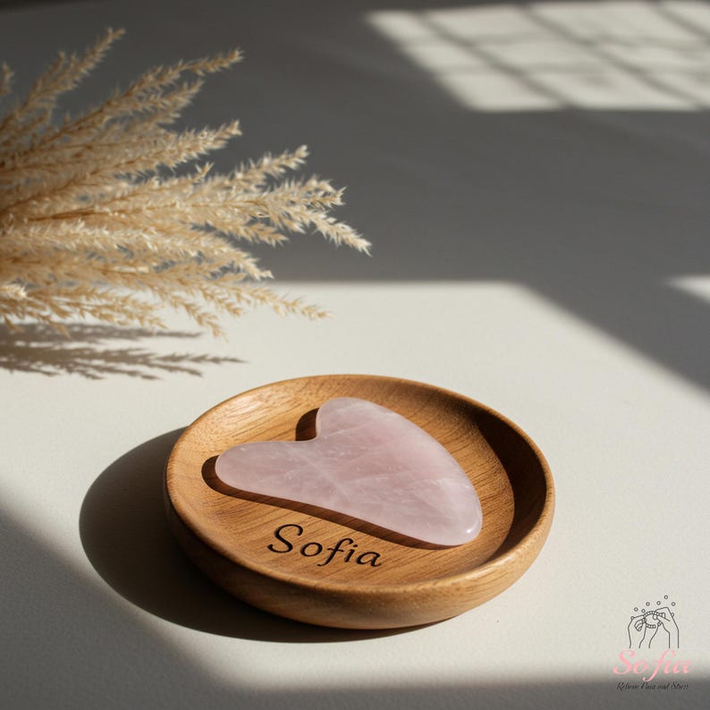 May include: A heart-shaped rose quartz gua sha tool sits on a small, round wooden dish. The dish is engraved with the name "Sofia". The gua sha is a pale pink colour, and the wood has a warm, natural tone. Dried ornamental grass is in the background.