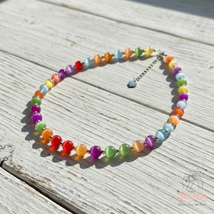 May include: A necklace featuring a circular arrangement of multicolored cat's eye beads in shades of red, orange, yellow, green, blue, and purple, interspersed with small silver-toned metal beads and finished with a silver-toned chain and heart charm.