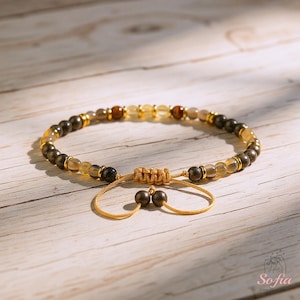 Puede incluir: Una pulsera de cuentas con una mezcla de cuentas doradas, marrones y transparentes. La pulsera tiene detalles dorados y un cierre de nudo corredizo. Las cuentas est&aacute;n ensartadas en un cord&oacute;n color canela, sobre una superficie de madera clara.
