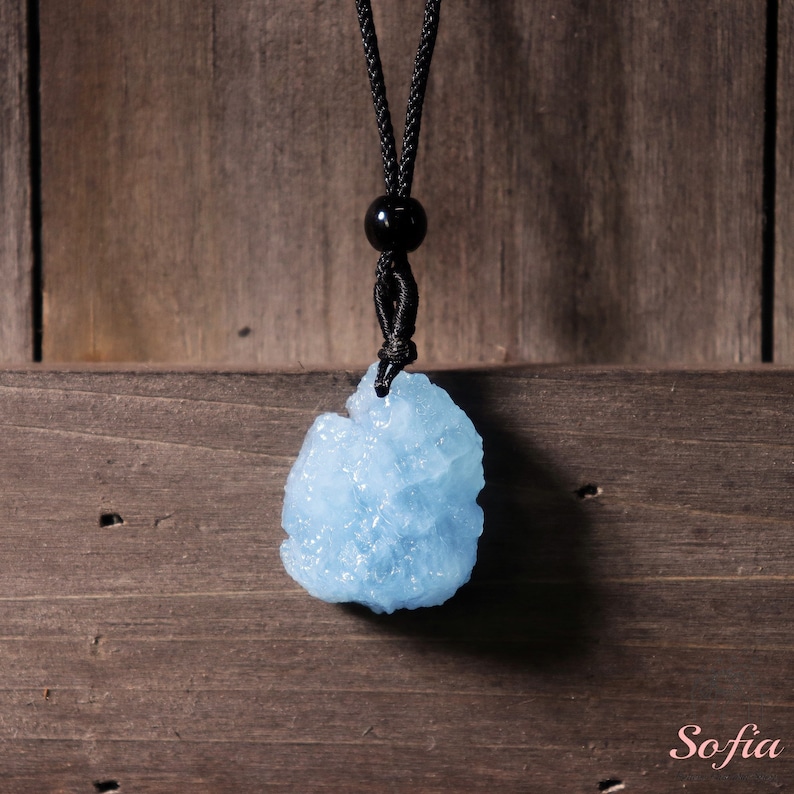 Puede incluir: Un colgante de cristal de aguamarina azul claro en un collar de cord&oacute;n negro. El colgante es un cristal natural de corte tosco con una forma &uacute;nica.