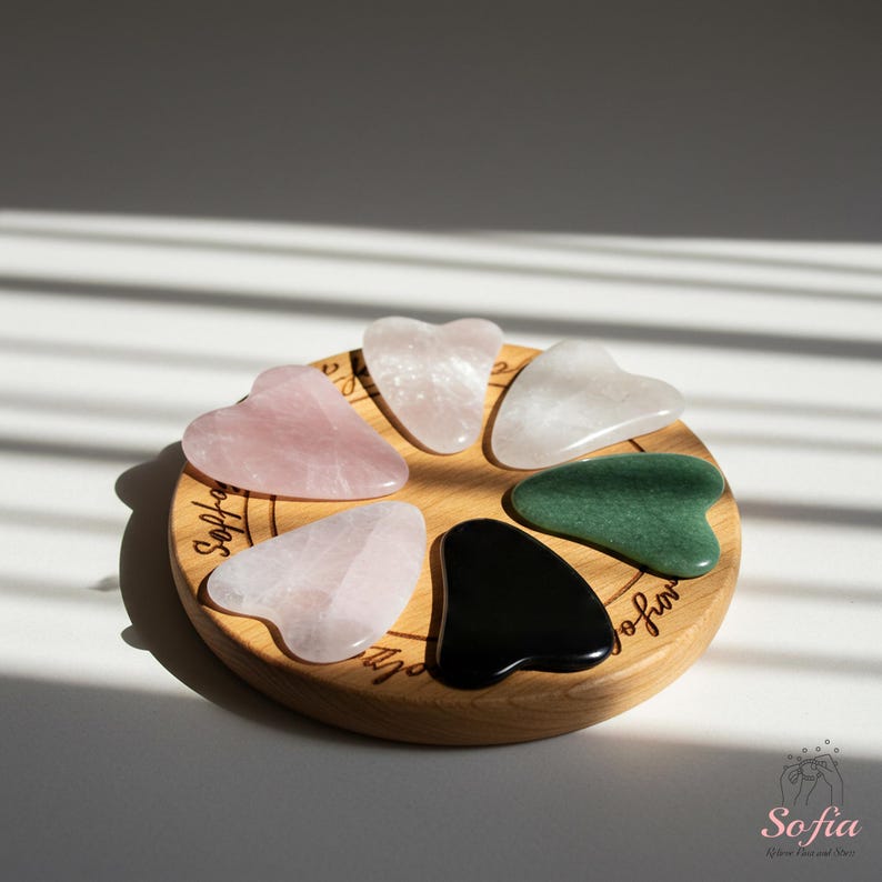 May include: A wooden display holds seven heart-shaped gua sha tools in various colors, including pink, white, green, and black. The tools are arranged in a circle, showcasing their different materials and textures. The display is on a white surface.