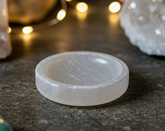 May include: A white selenite crystal dish with a smooth, polished surface. The dish is round and has a slight lip around the edge. It is perfect for holding small items, such as jewelry or crystals.