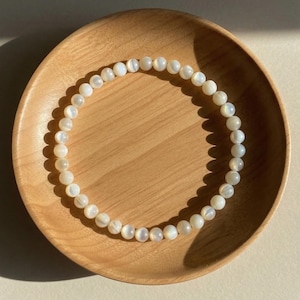 May include: A circular bracelet composed of small, smooth, off-white spherical beads with a subtle pearlescent sheen. The beads are strung together in a continuous loop, creating a simple and uniform design that reflects light across their surfaces.