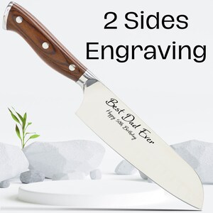 Puede incluir: Un cuchillo de chef de acero inoxidable con mango de madera. El cuchillo tiene grabado el texto "Best Dad Ever" y "Happy 50th Birthday". El cuchillo est&aacute; sobre una superficie blanca con piedras y una peque&ntilde;a planta.