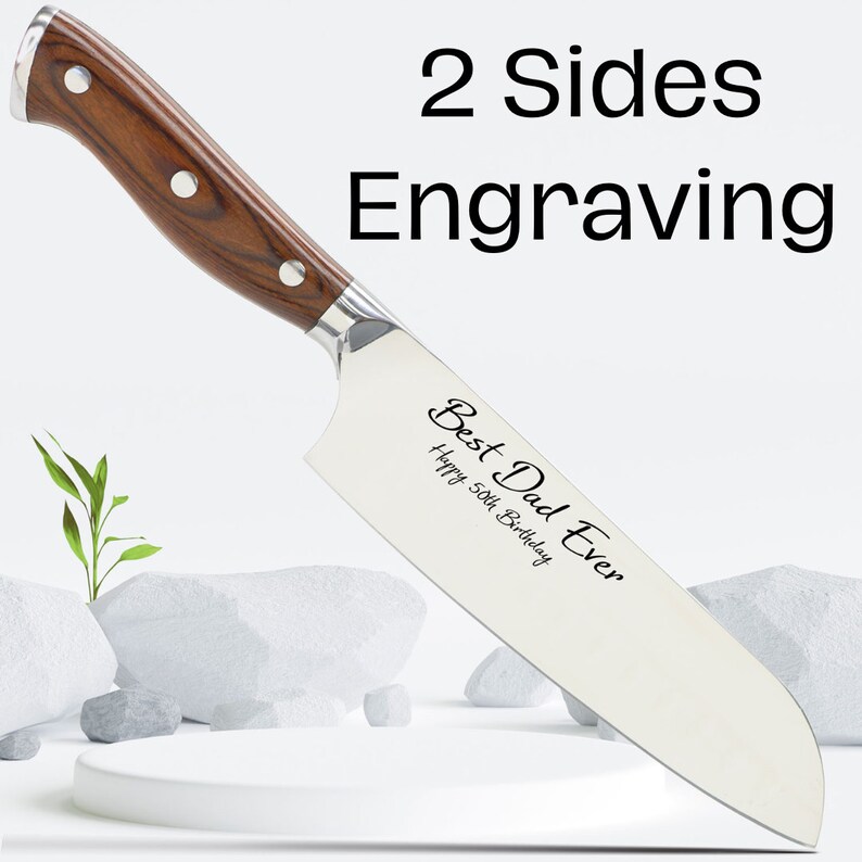 Puede incluir: Un cuchillo de chef de acero inoxidable con mango de madera. El cuchillo tiene grabado el texto "Best Dad Ever" y "Happy 50th Birthday". El cuchillo est&aacute; sobre una superficie blanca con piedras y una peque&ntilde;a planta.