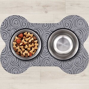 May include: A gray bone-shaped pet food mat with a swirling pattern. Two stainless steel bowls are on the mat, one filled with dog food and the other empty.